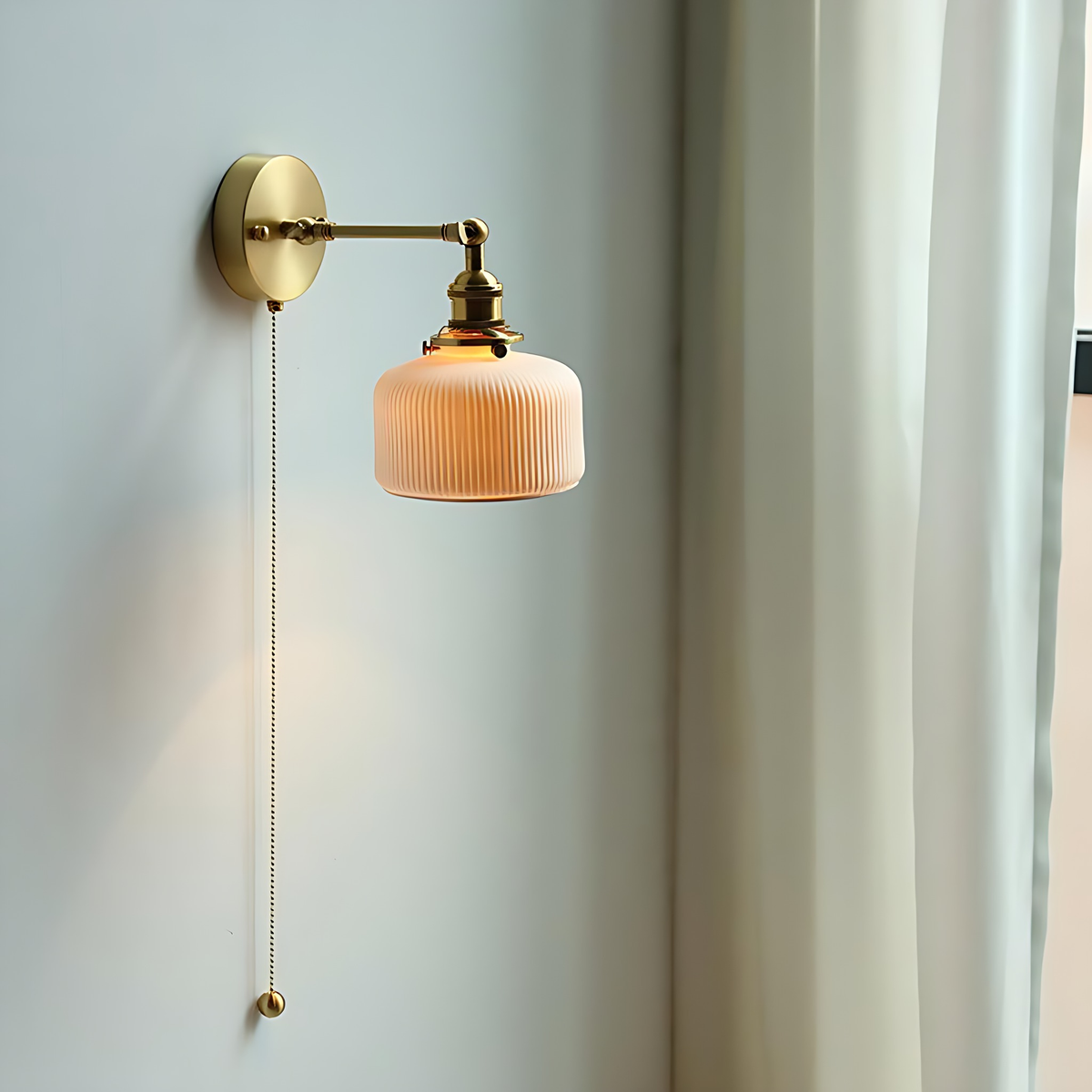 Fia wall sconce lit on a pale wall, its brass bead pull chain hanging down
