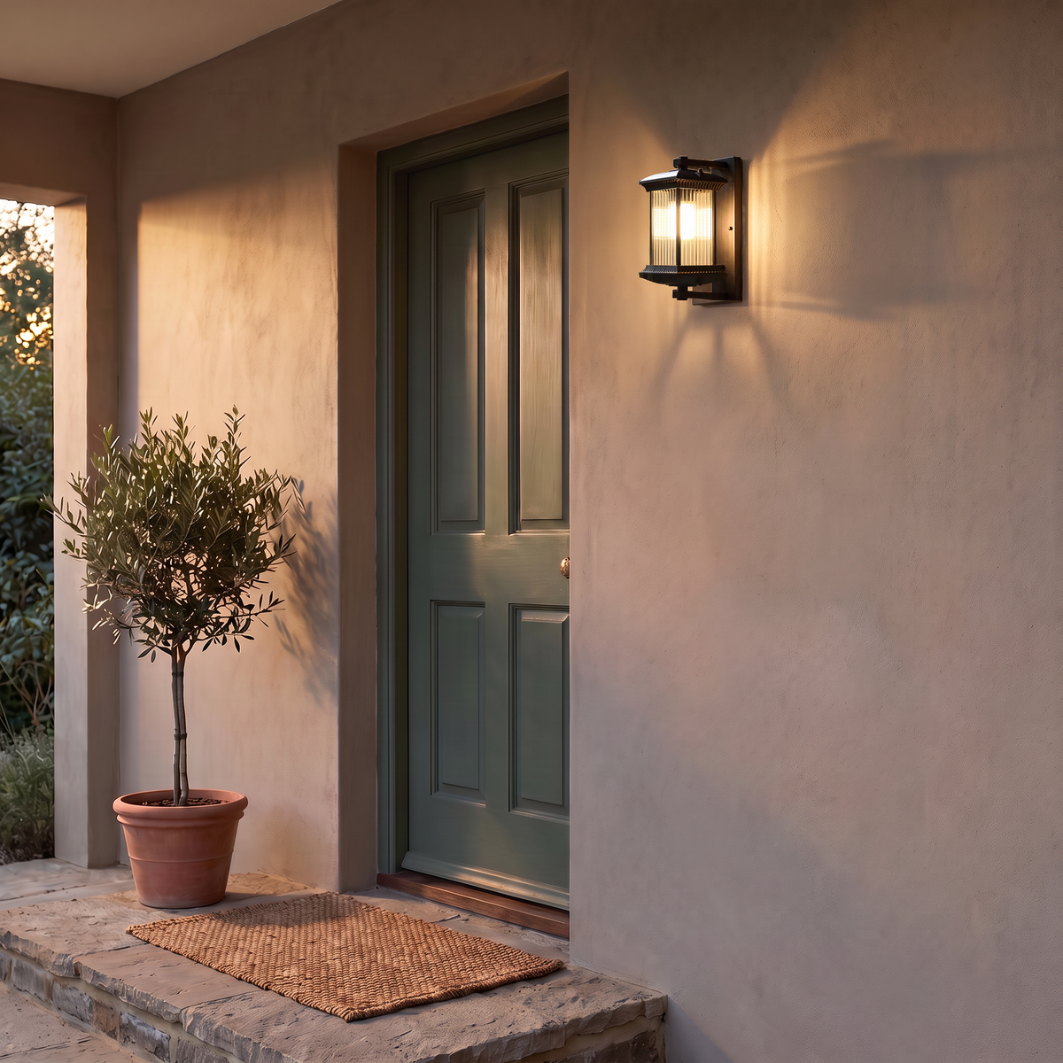 Black outdoor wall lantern lit beside a green front door on a stucco porch at dusk, with an olive tree in a terracotta pot