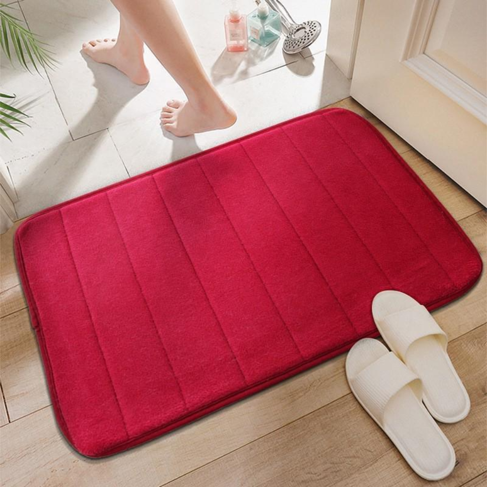 Red microfiber bath mat on a tile floor beside white slippers