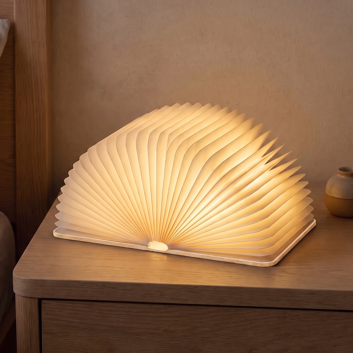 Sula book lamp with light wood covers, opened flat into a glowing fan on an oak nightstand