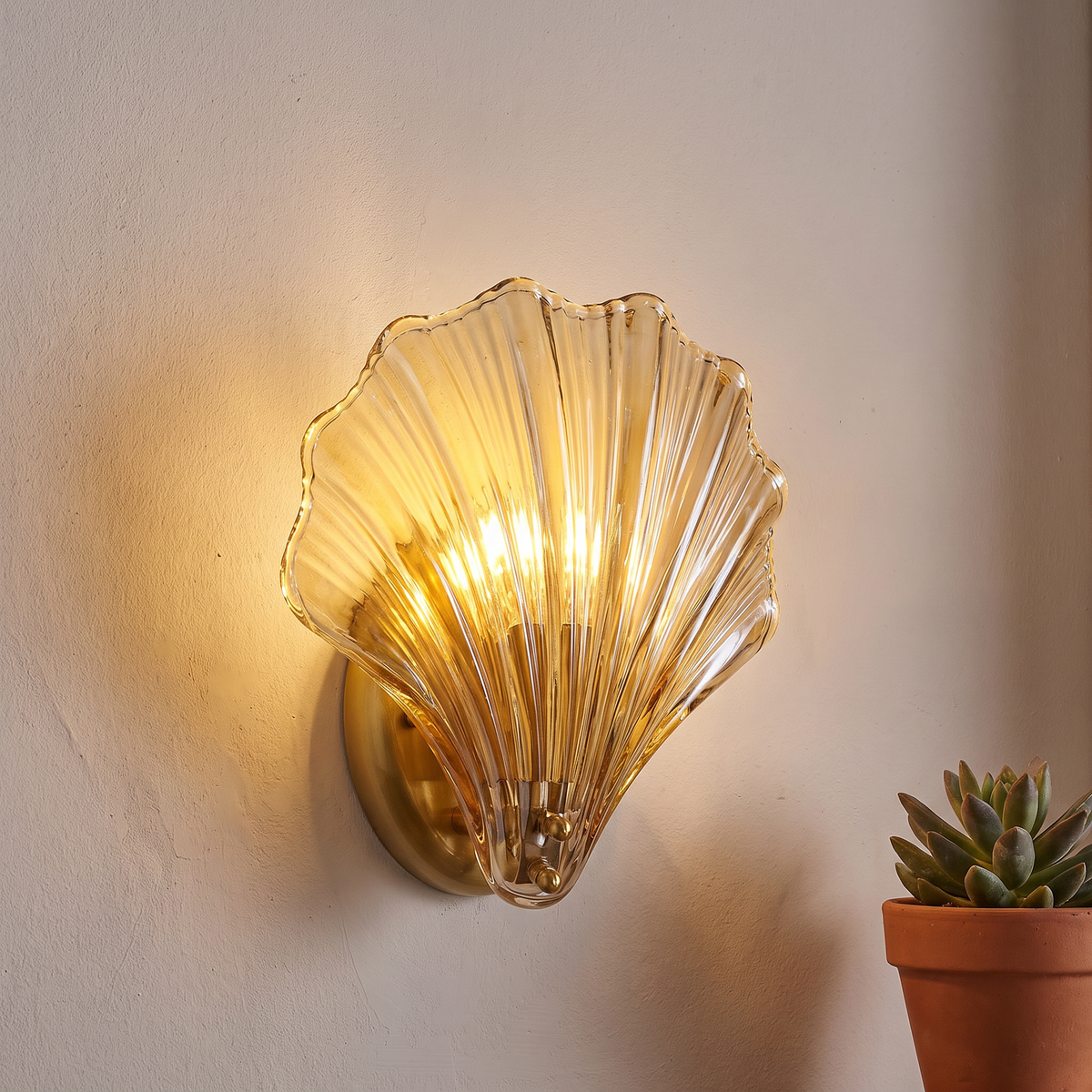 Close view of the Tyra sconce glowing on a plaster wall beside a small potted plant