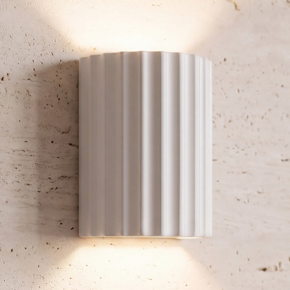 Unni LED wall sconce, white fluted shell, lit up and down on a travertine wall