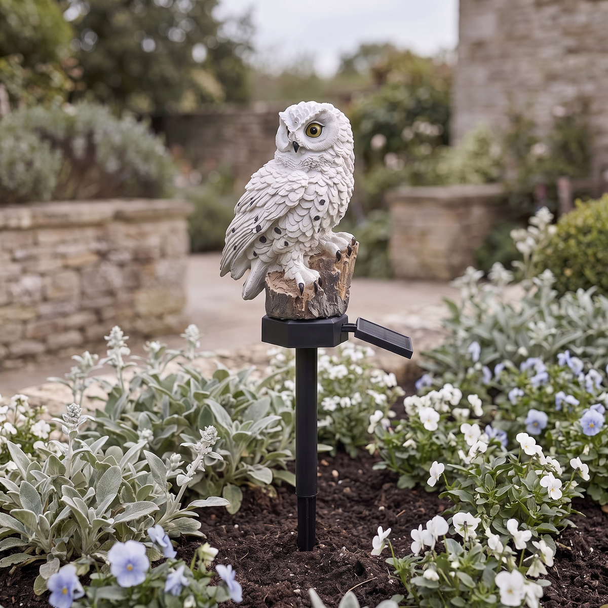 Alta solar owl light staked into a flower bed in daylight, solar panel beside the base