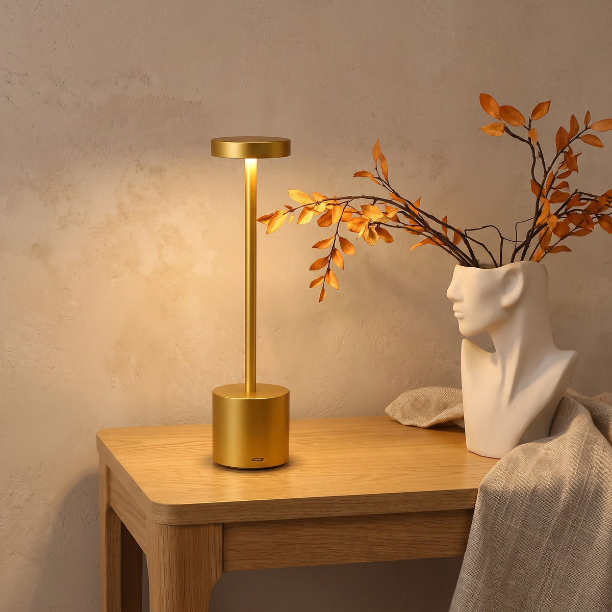 Gold Asta cordless table lamp lit beside a vase of leaves