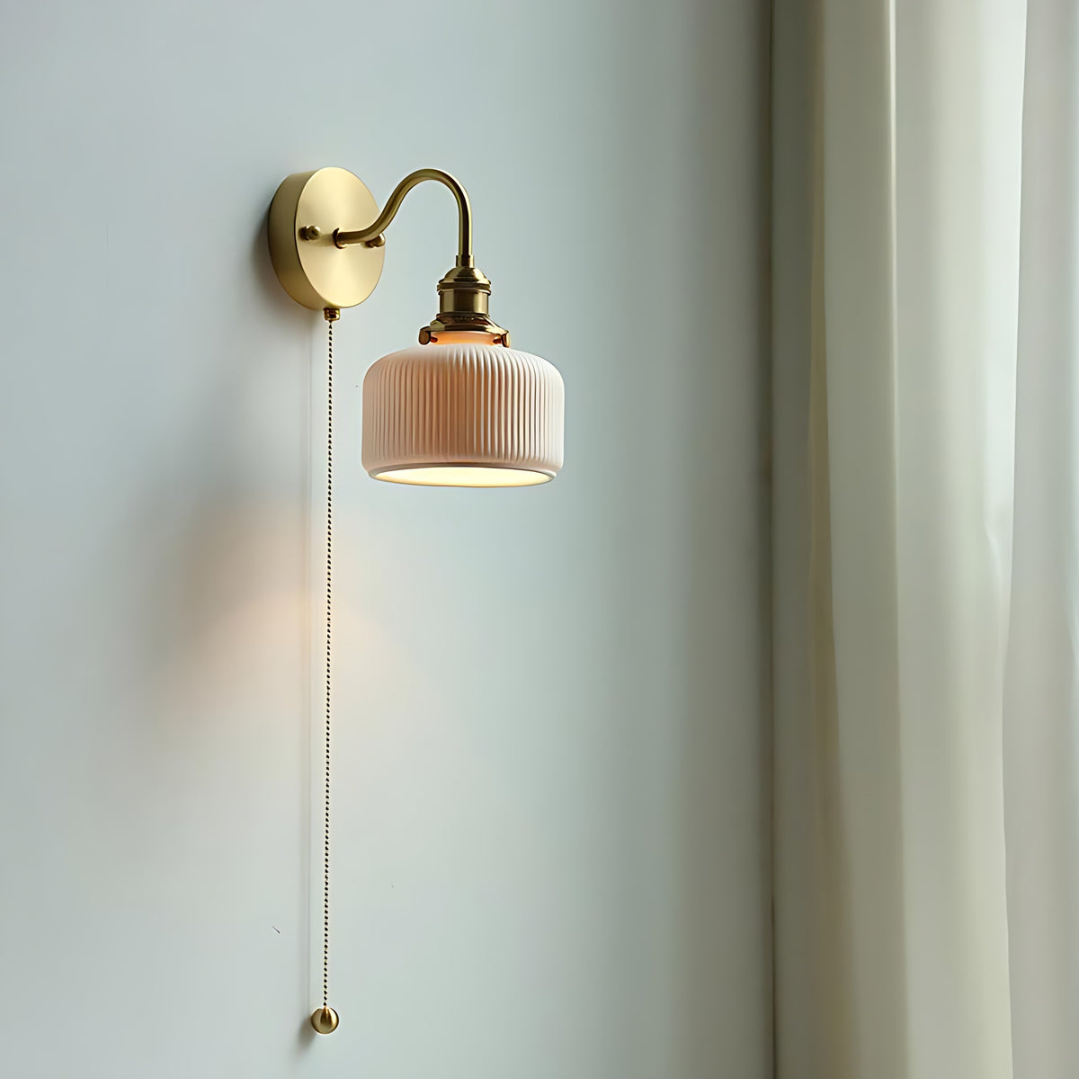 Brass wall sconce with a curved arm and ribbed ceramic shade, lit, on a pale wall beside a sheer curtain