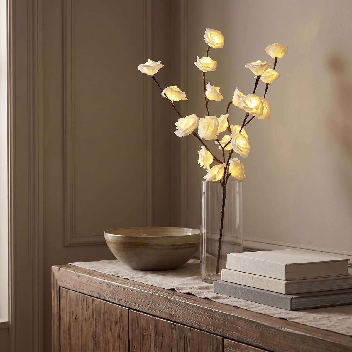 Lighted rose branches in a tall glass vase on a sideboard