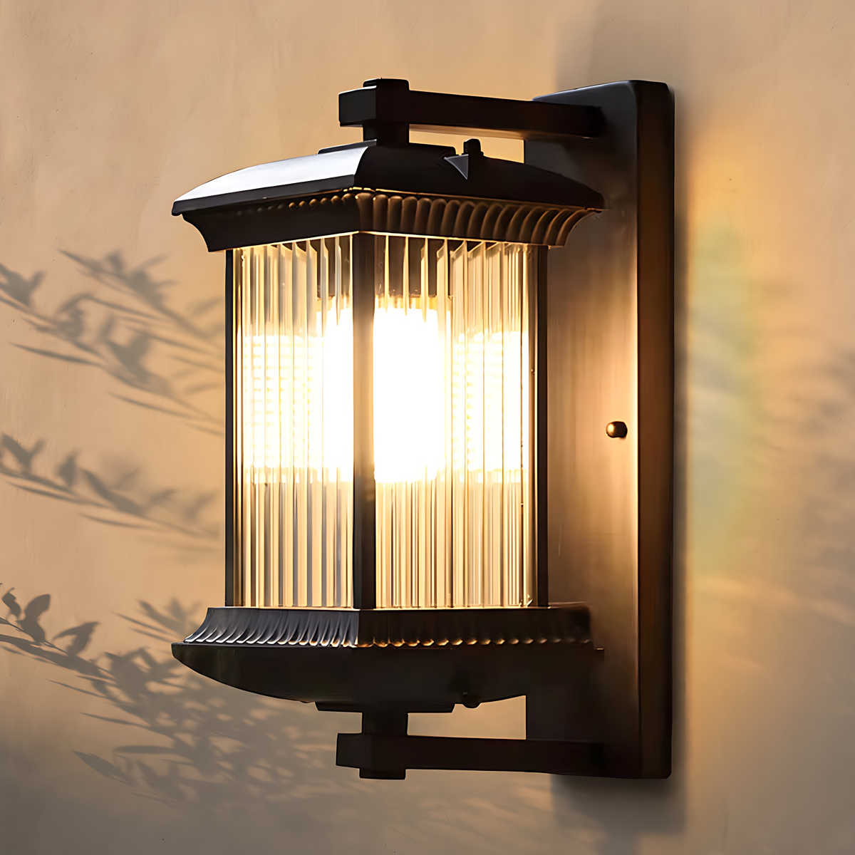 Meja outdoor wall lantern in black with clear fluted glass, lit, on a warm stucco wall with leaf shadows