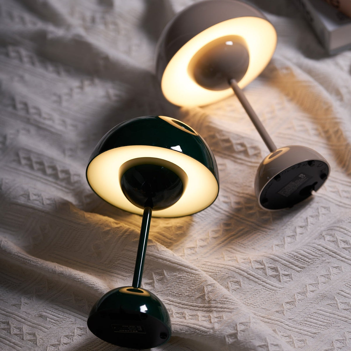Two Oona table lamps lying on a bed, lit, showing the glowing underside of the shade