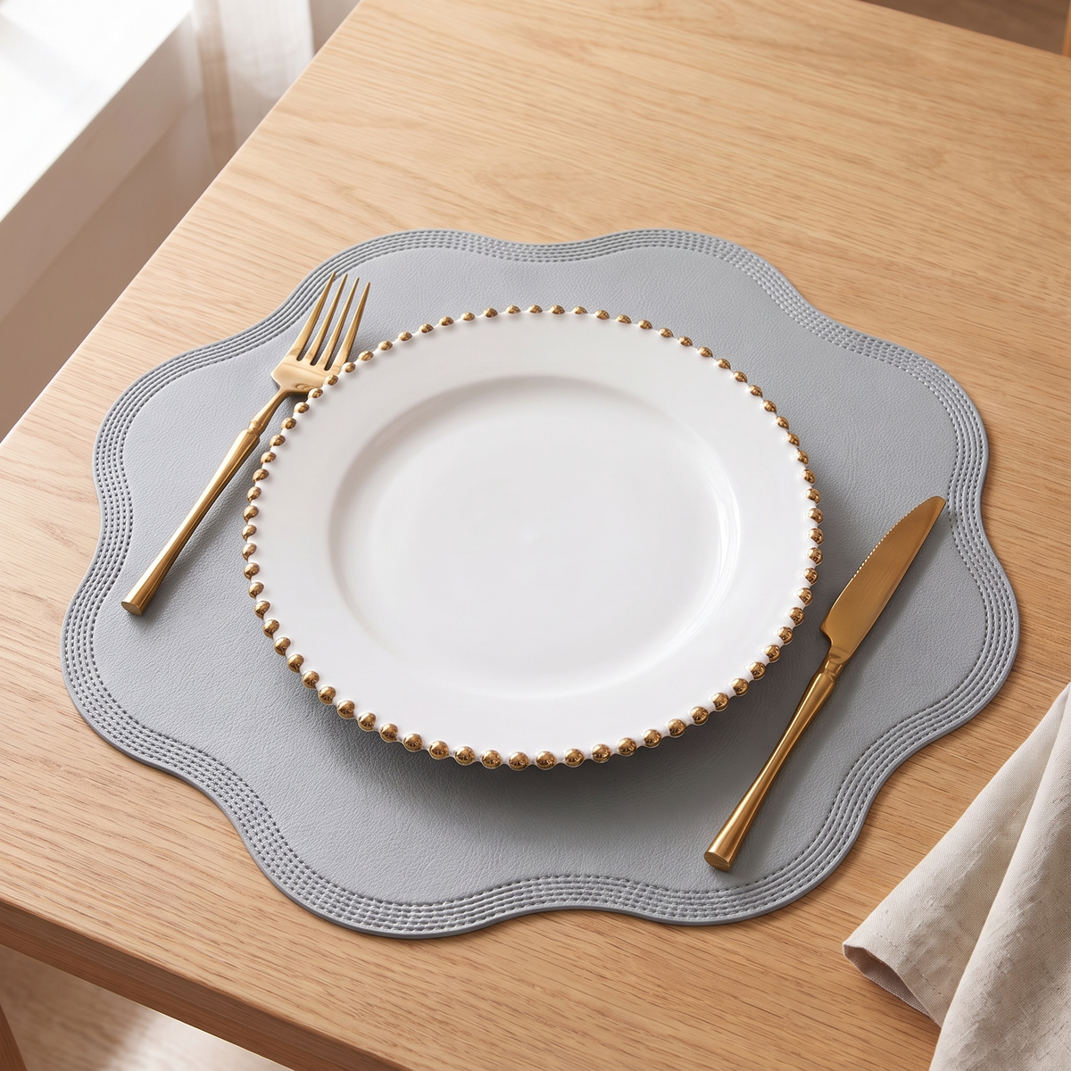 Scalloped placemat in gray with a white plate and gold cutlery on an oak table