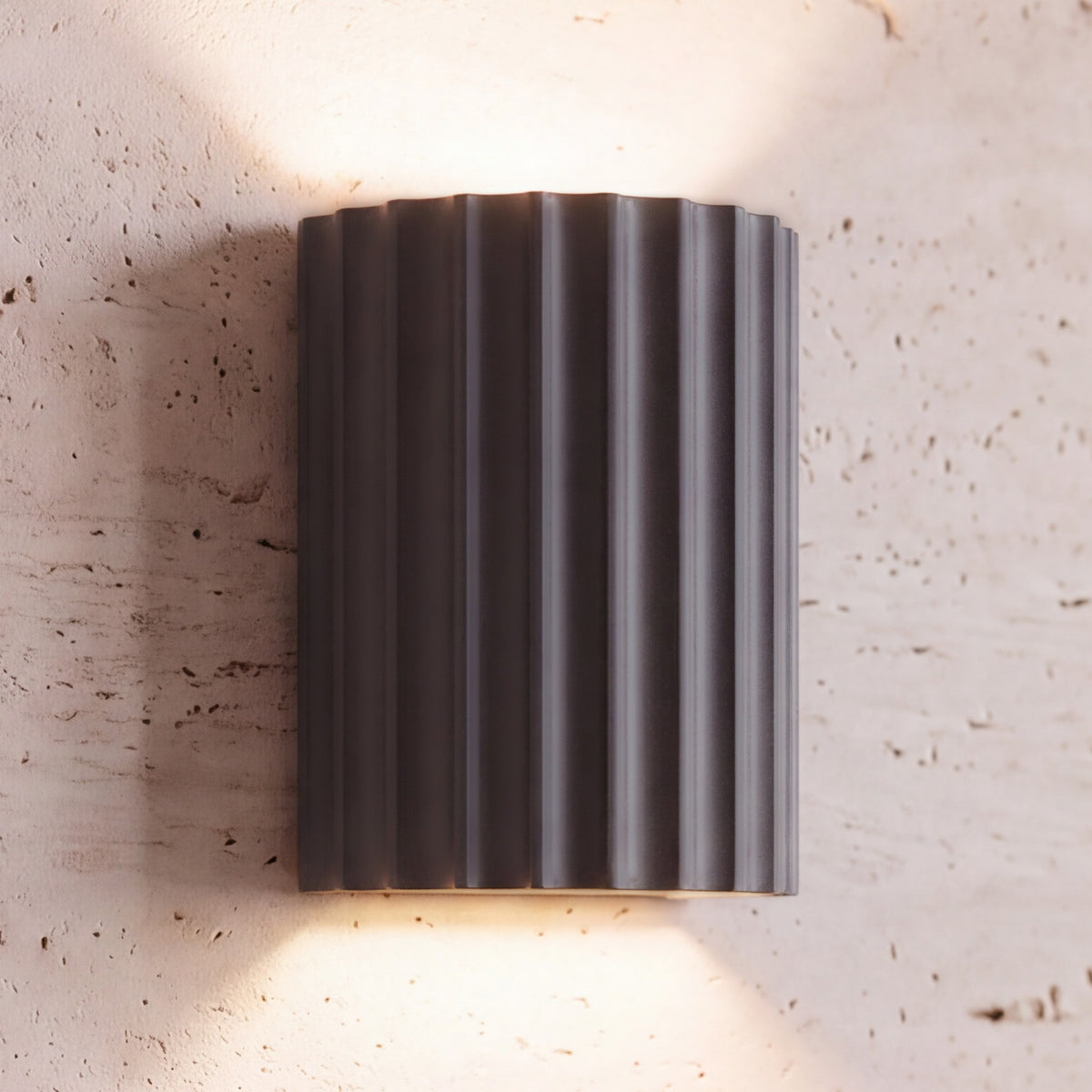 Unni LED wall sconce, gray fluted shell, lit up and down on a travertine wall